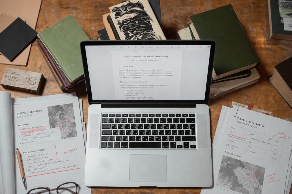 Laptop open to a country research document on a wooden desk surrounded by annotated source notes and books